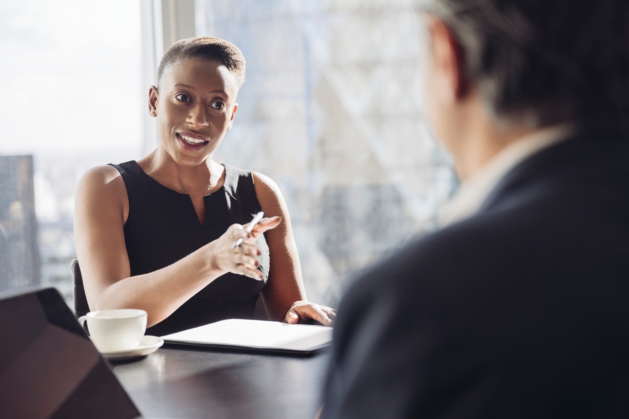 Over the shoulder view of well-dressed British executive in mid 30s sitting at conference table in sunny meeting room and smiling as she makes her point.